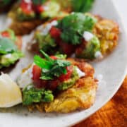 A plate of traditional fried green plantains topped with guacamole, salsa, and a dollop of cream garnished with cilantro, accompanied by lemon wedges on the side.
