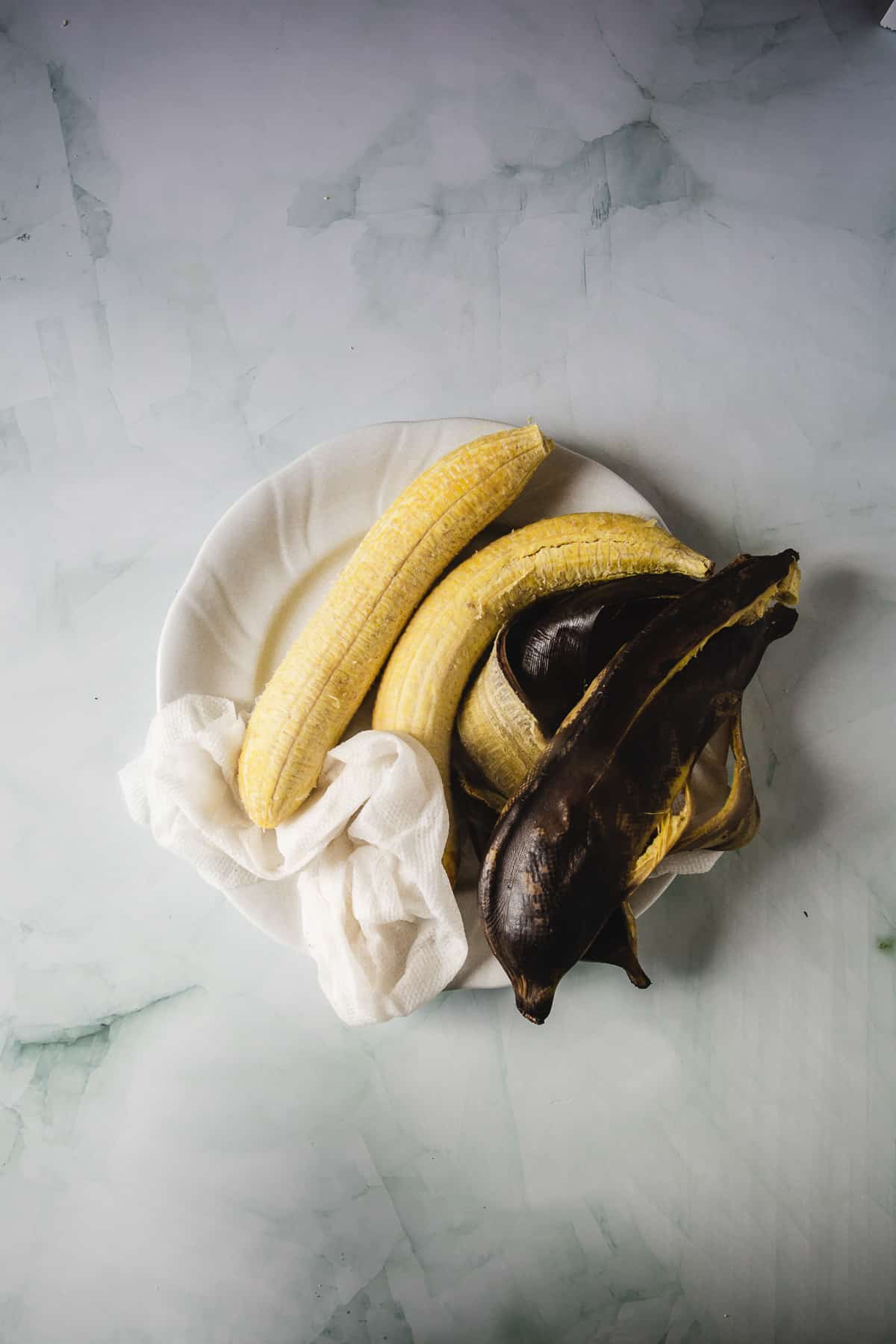 A bowl containing one whole plantain, one partially-peeled banana, and one completely-peeled plantain, displayed on a marbled surface next to Colombian Patacones.