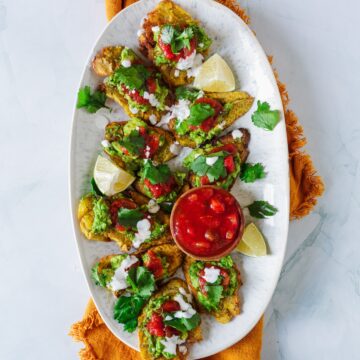 A platter of freshly prepared colombian patacones garnished with lime wedges, cilantro, and salsa on a white surface.