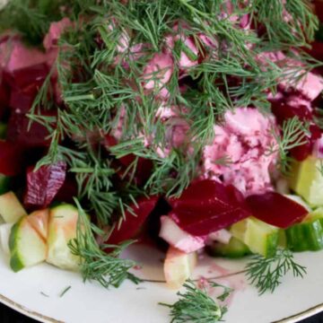 A white plate with a beet and cucumber salad is topped with a generous amount of fresh dill. Perfect for those seeking quick recipes, the salad includes diced beets and cucumbers, mixed with a creamy pink dressing. A spoon rests on the side of the plate.