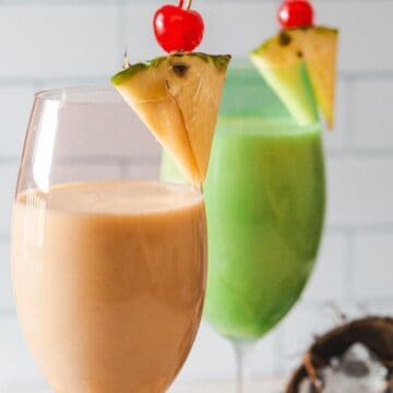 Two must-try colorful tropical drinks, one orange and one green, garnished with pineapple slices and cherries, are presented against a white tile background.