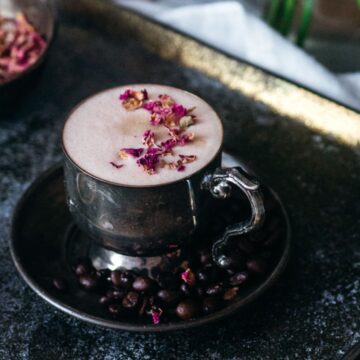 A cup of frothy beverage garnished with pink petals on a saucer surrounded by coffee beans, with cardamom pods and dried rose petals in the background, all presented on a moody,