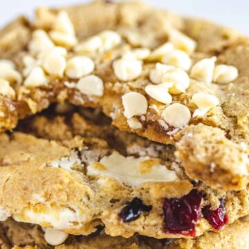 Close-up image of a stack of cookies, featuring white chocolate chips and pieces of cranberries. The top cookie has a bite taken out of it, revealing a chewy texture inside. These dessert classics have a light golden-brown color and a slightly crumbly surface, making them hard to forget.