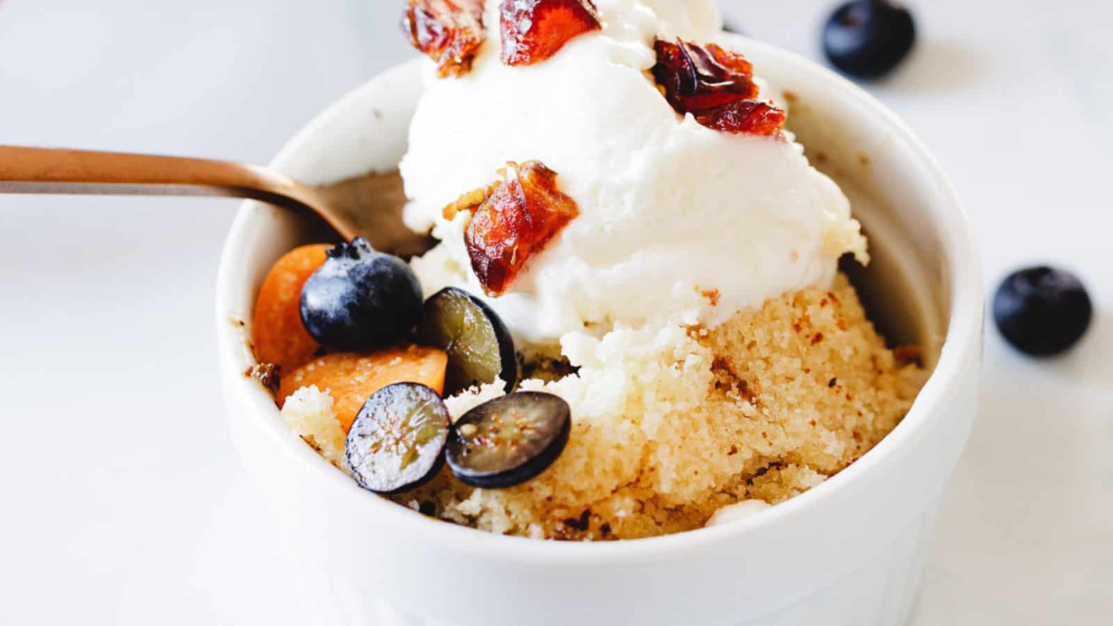 A close-up of a bowl of couscous topped with a dollop of whipped cream, fresh blueberries, and caramelized dates. Remember This? A spoon is scooping a portion from the bowl, and a few blueberries are scattered on the white surface around the bowl.