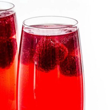 Two glasses of red raspberry drink with whole raspberries floating inside, set against a bright white background.