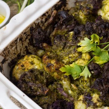A white baking dish filled with roasted cauliflower florets covered in a green herb mixture, just like Grandma's recipes, garnished with a sprig of parsley. A small white cup containing yellow dressing is placed to the side, and green onions are scattered around.
