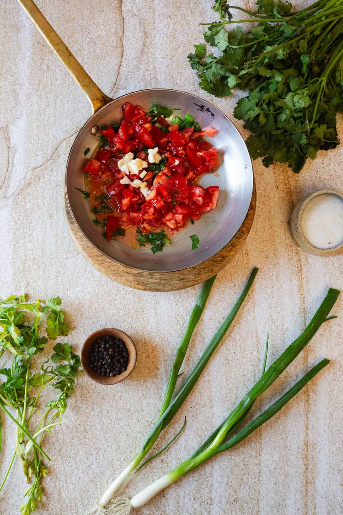 A preparation of diced tomatoes and garlic in a metal bowl with a wooden handle, accompanied by fresh herbs and spices on a kitchen countertop.