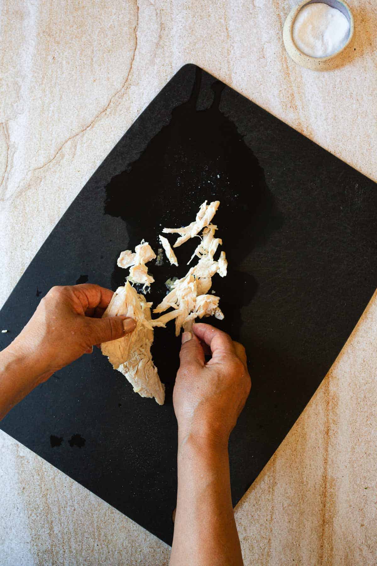 A person is shredding chicken on a black cutting board with a small dish of salt to the side.