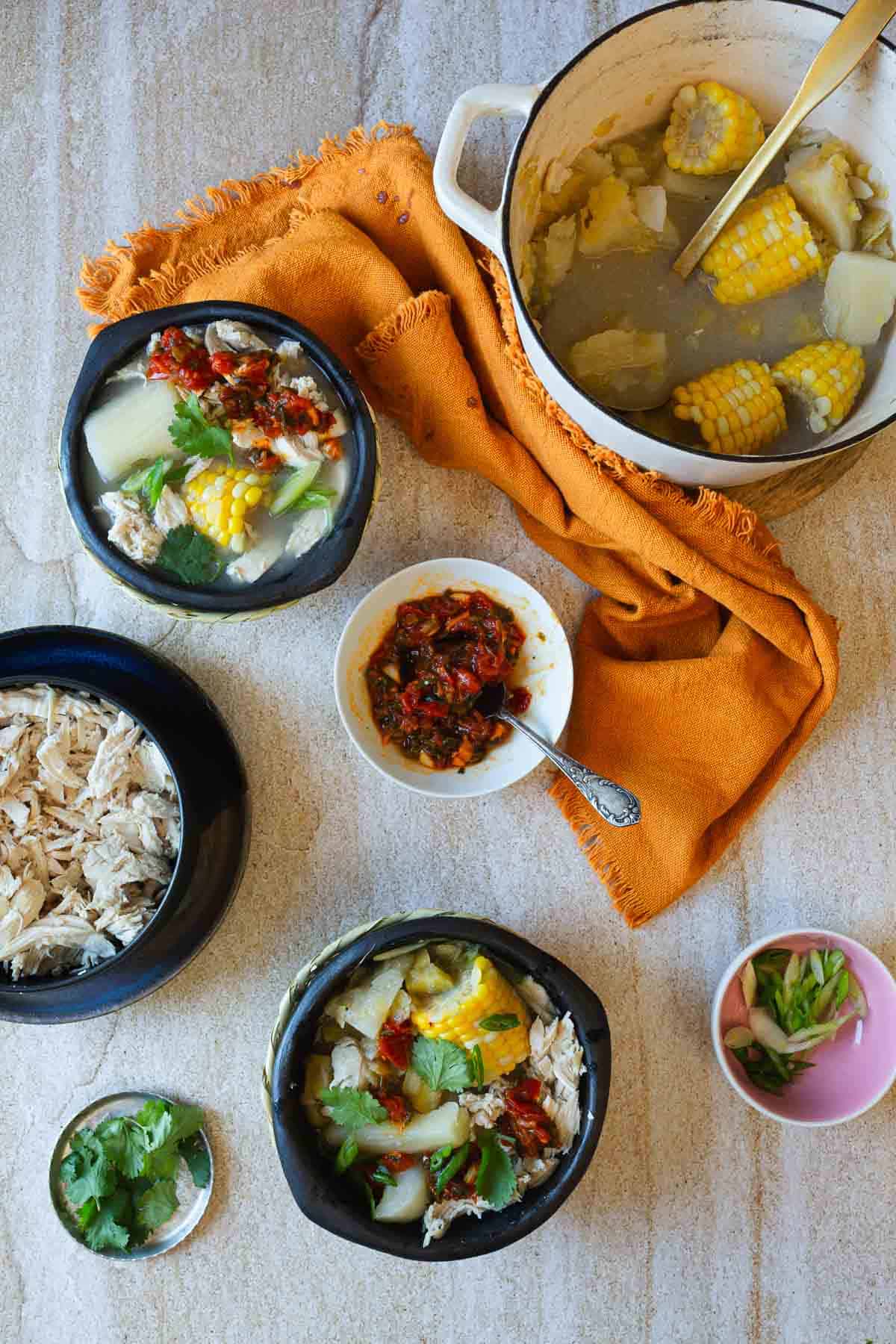 A flat lay photograph of a meal setup with two bowls of chicken soup, a side dish of salsa, shredded chicken on a plate, a pot containing corn cobs and ladle, and small bowls of herbs and onions, all arranged on a table with an orange cloth napkin.