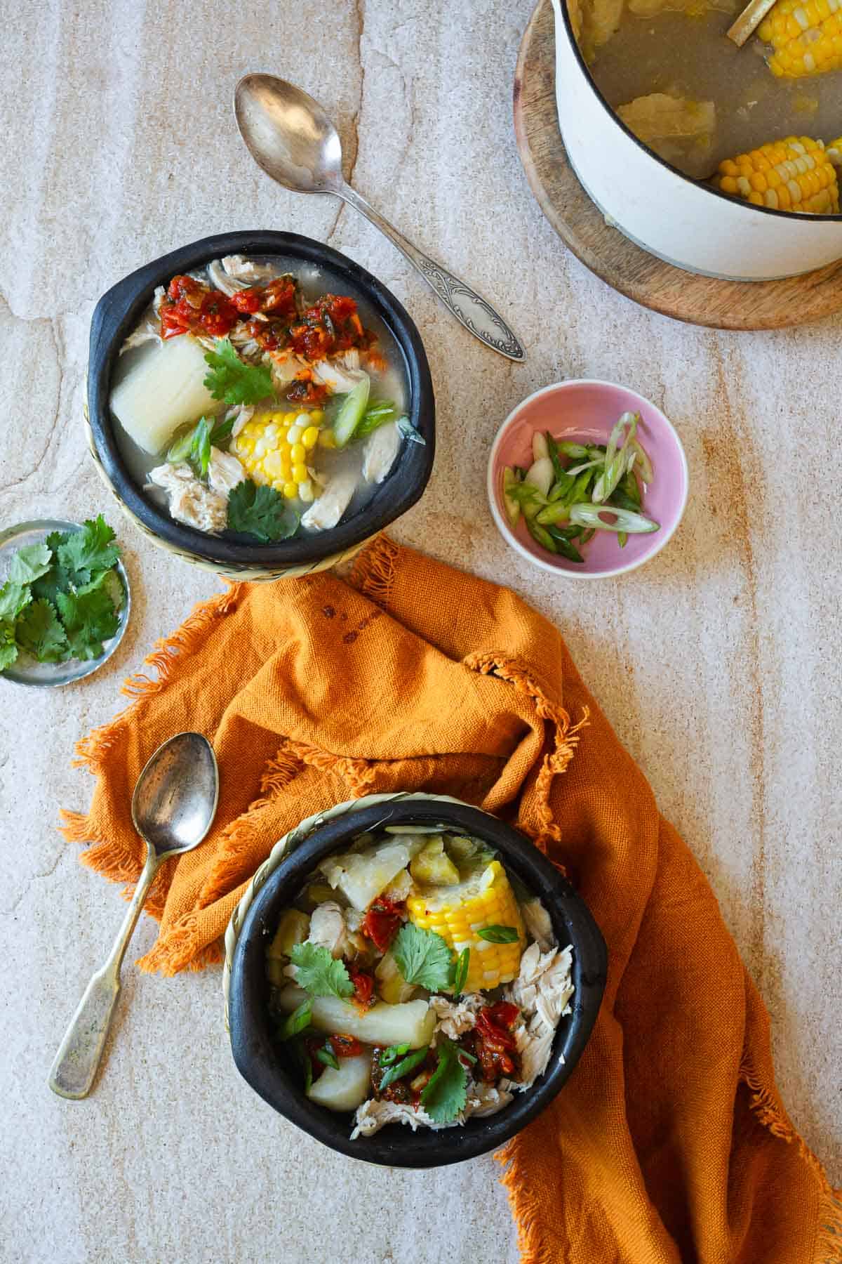 Two bowls of chicken sancocho with corn on a rustic surface, accompanied by a bowl of sliced scallions and a pot with more soup in the background.