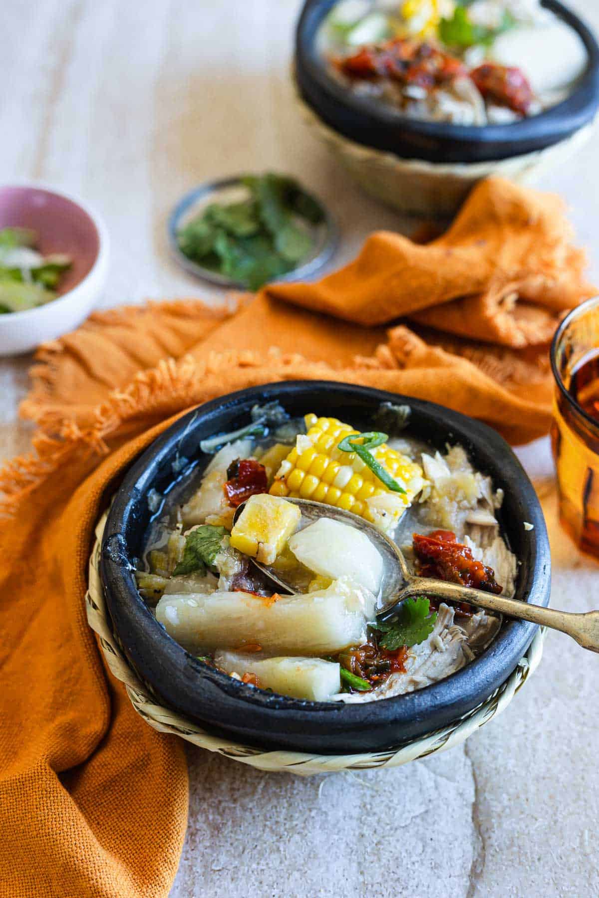 A bowl of chicken soup with various vegetables, including corn on the cob and leafy greens, garnished with herbs, accompanied by small side dishes and a spoon, served on a textured table with a cloth napkin.