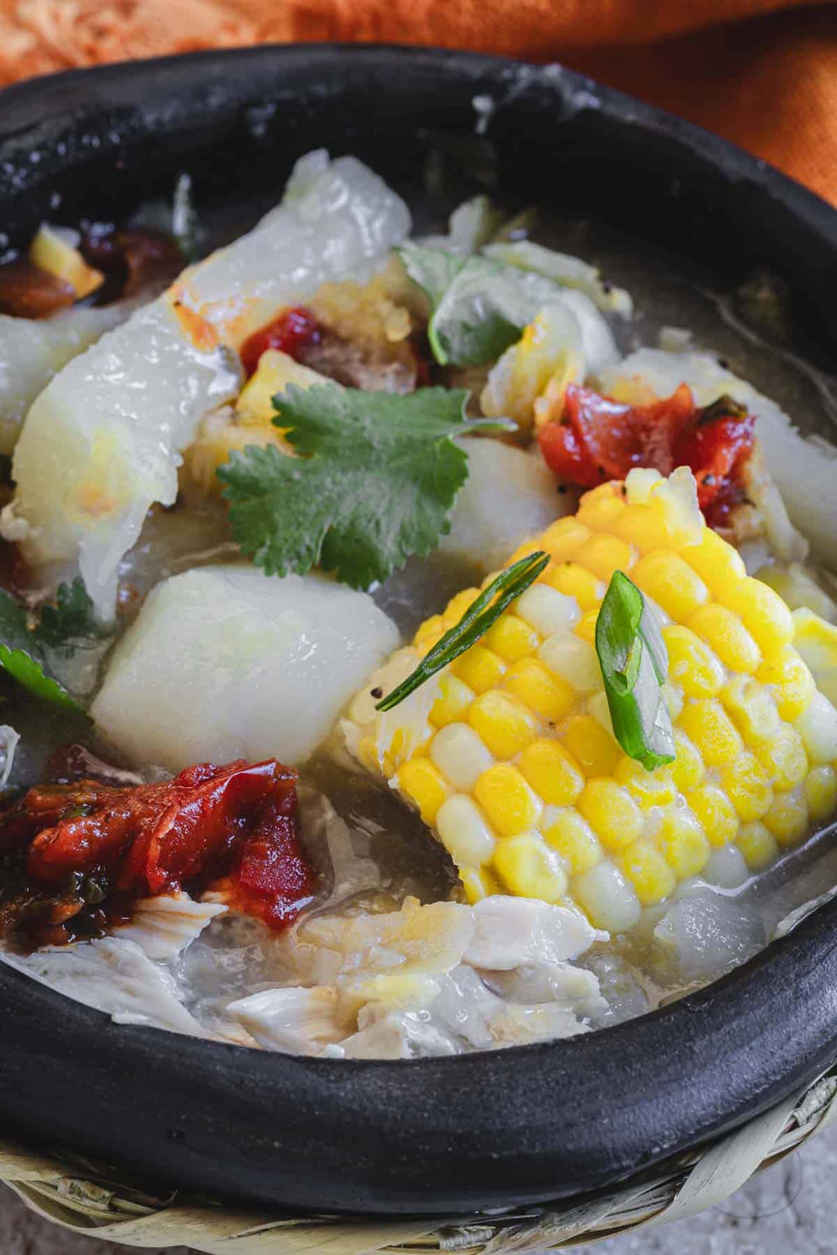 A colombian chicken sancocho stew served in a black stone pot, garnished with corn, cilantro, and red peppers.