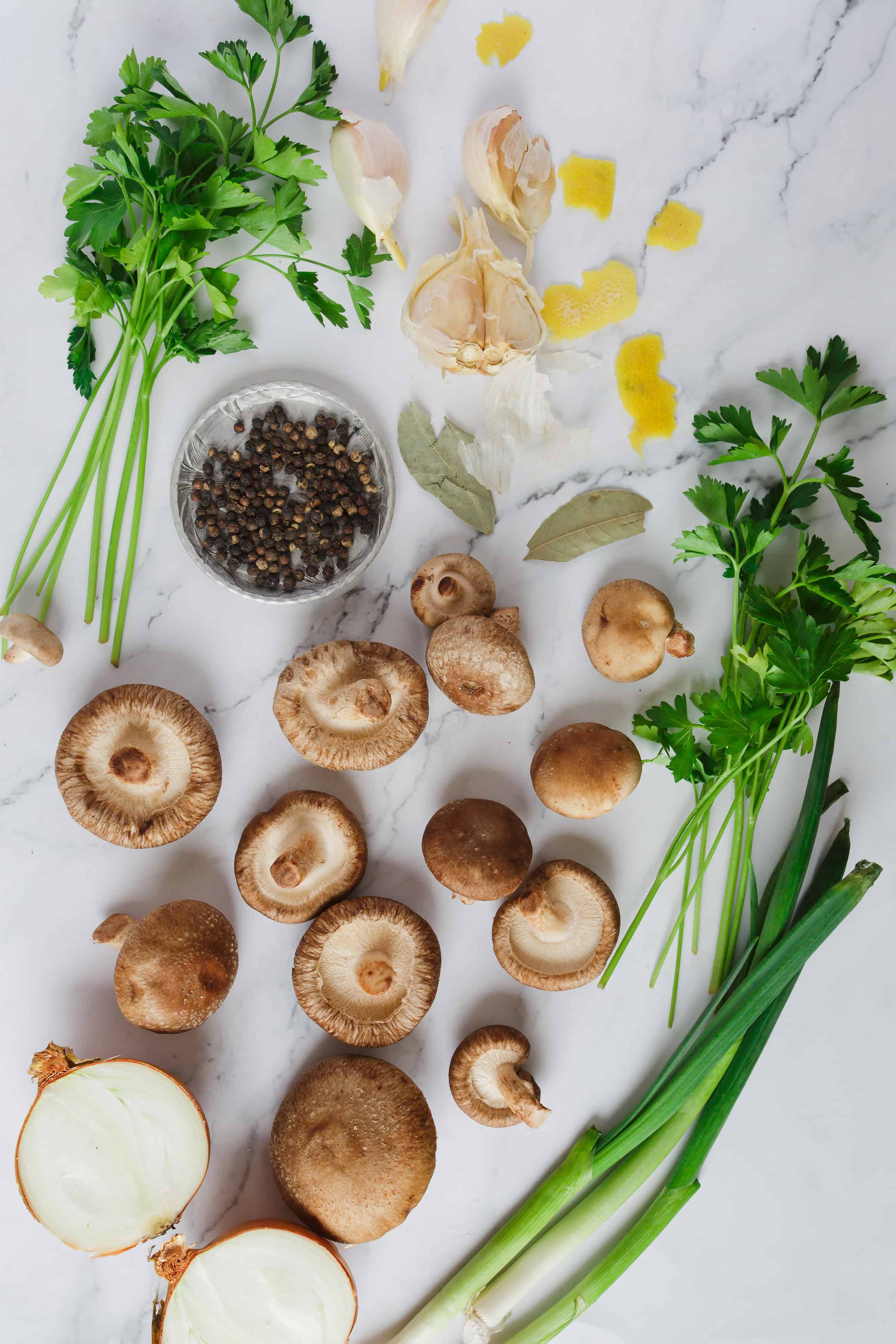 Various fresh ingredients arranged neatly on a marble surface including shiitake mushrooms, garlic cloves, green onions, parsley, an onion halved, peppercorns, and lemon zest.