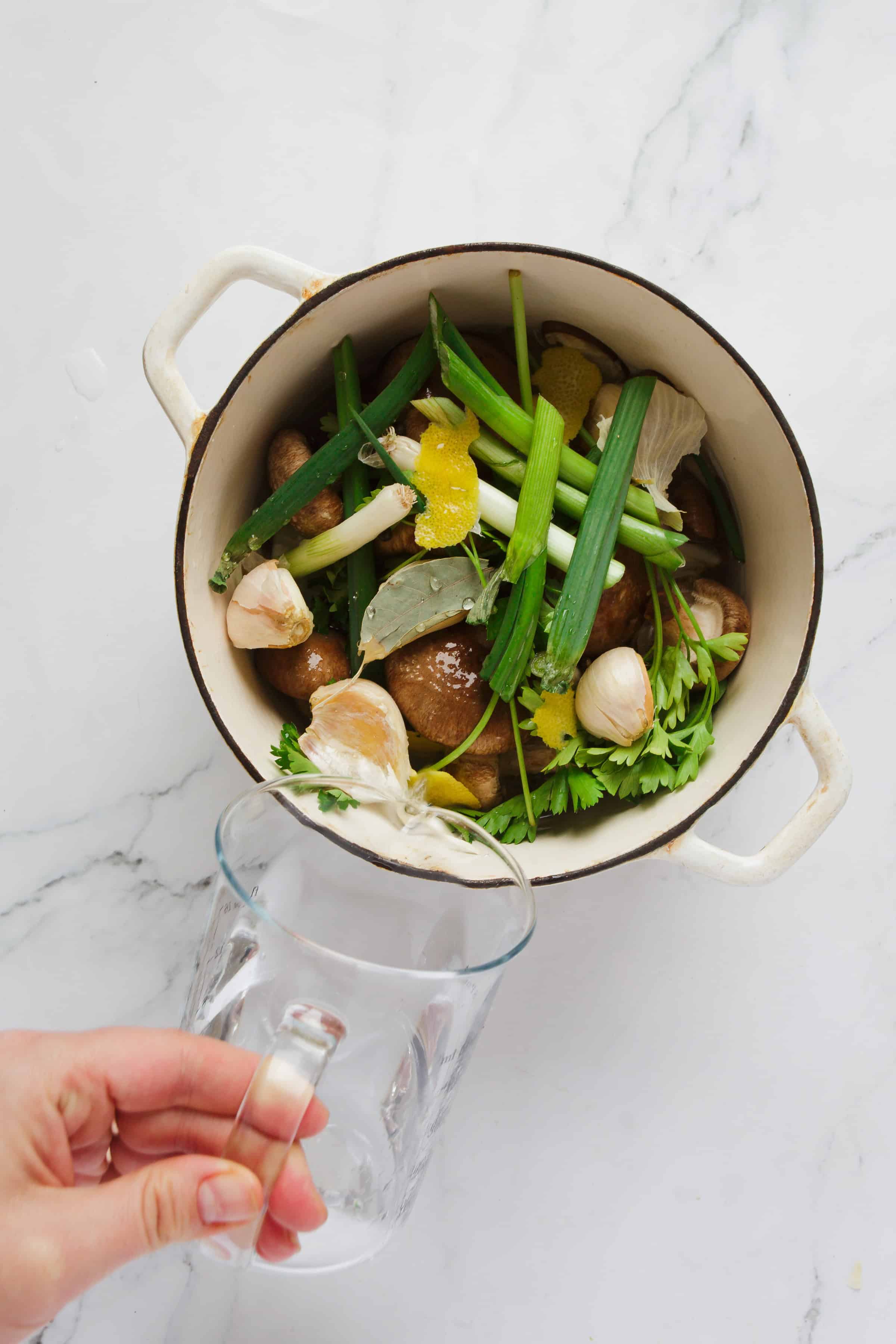 A white enameled pot filled with fresh vegetables, including green onions and garlic, on a marble surface.