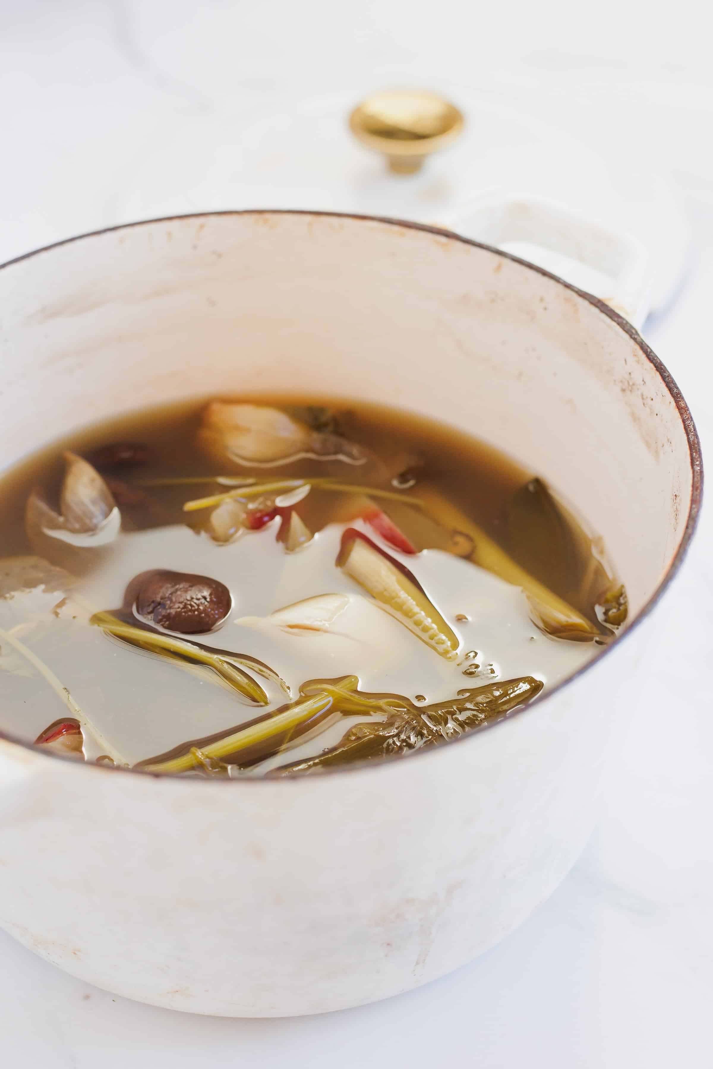 A large, white enameled pot containing a vegan dashi with various ingredients including whole garlic cloves, sprigs of herbs, shiitake mushrooms, and spices on a white surface.