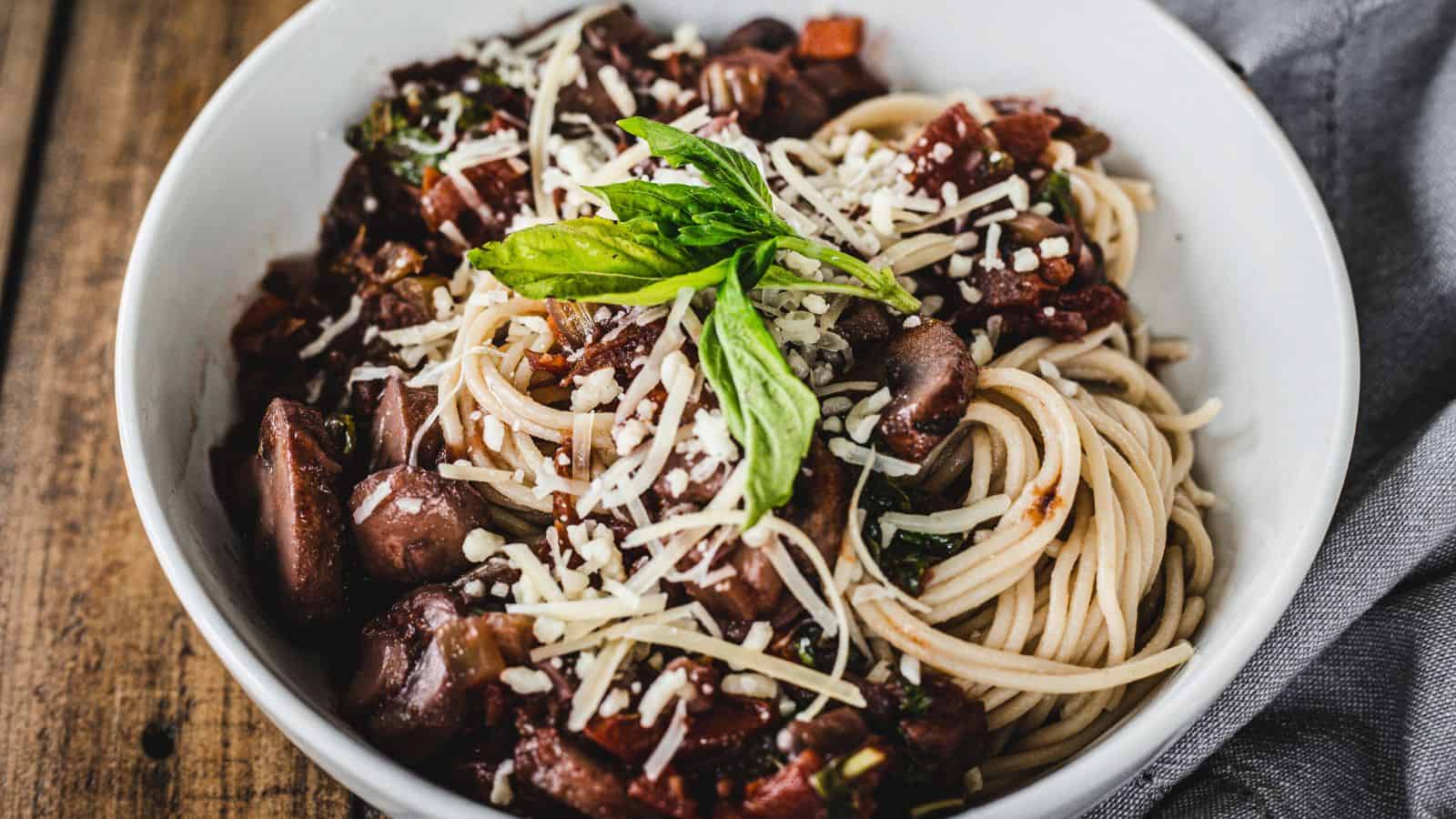 A bowl of spaghetti topped with a rich, chunky tomato sauce with diced vegetables and olives, sprinkled with grated cheese and garnished with fresh basil leaves, featured in our weekly food guide.