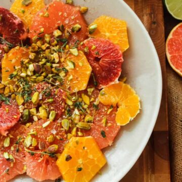 A vibrant citrus salad featuring slices of orange, grapefruit, and blood orange from Grandma's recipes, garnished with chopped pistachios on a white plate, with whole citrus fruits and a lime half visible in the background.
