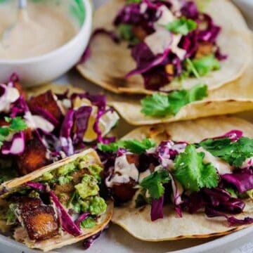 Three quick fish tacos served on a gray plate, garnished with red cabbage, cilantro, and lime wedges, accompanied by a side of creamy sauce.