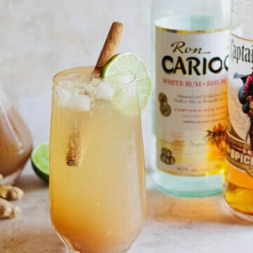 A cocktail glass filled with a light brown drink containing ice, a lime wheel, and a cinnamon stick is on a table. Behind the glass are bottles of Ron Carioca white rum and Captain Morgan spiced rum, along with a carafe of a brown liquid and pieces of fresh ginger—truly one of the must-try drinks.