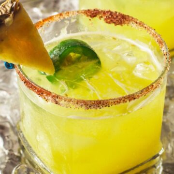 A close-up of a chilled pineapple margarita cocktail with a salt and chili powder rim, garnished with a slice of lime and a pineapple wedge, served on a bed of crushed ice. You