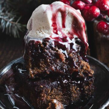 A serving of warm pudding topped with a scoop of melting vanilla ice cream and a drizzle of red berry sauce, presented on a rustic wooden table alongside festive pine branches and red berries.
