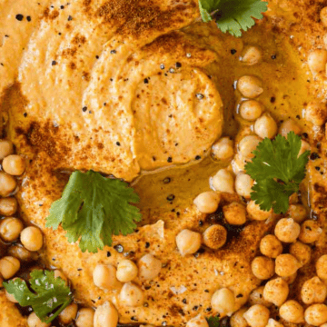 A bowl of hummus topped with whole chickpeas, paprika, black pepper, and fresh cilantro leaves. The smooth, creamy texture with a swirl pattern in the center makes it both inviting and delightful for those seeking quick recipes. The dish is set on a light-colored surface.