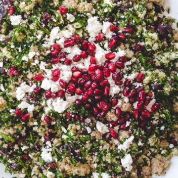 A white plate filled with a couscous salad, garnished with chopped herbs, olives, crumbled feta cheese, and a central topping of pomegranate seeds. Perfect for those seeking quick recipes, a metal spoon and a beige cloth napkin are placed to the side.