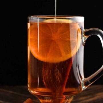 A glass mug of tea with a lemon slice, cinnamon stick, and honey being poured in—an inviting addition to any brunch drinks spread, set against a dark background.