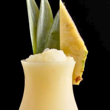 A frosty pineapple cocktail served in a tall glass, garnished with a slice of pineapple and a leafy pineapple top, against a dark background.