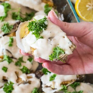 A close-up of a hand holding a baked stuffed clam topped with creamy sauce and garnished with fresh dill. In the background, the glass dish of additional clams is visible on this Monday evening