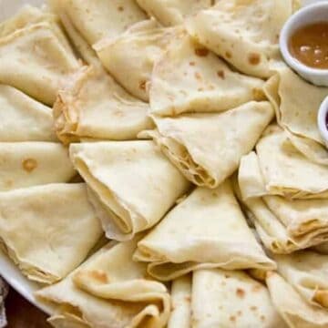 A plate of neatly folded crepes accompanied by small bowls of apricot and strawberry jam, set on a table with a lace cloth, perfect for quick recipes.