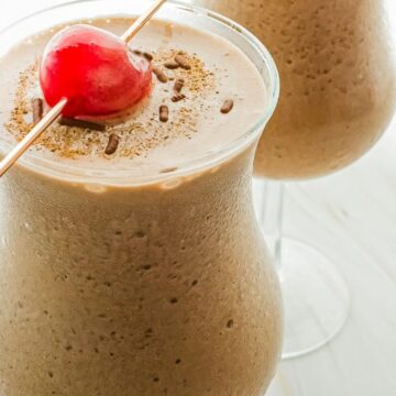 Two glasses of chocolate milkshake topped with whipped cream and garnished with a red cherry on a white tabletop. the milkshakes are frothy and served with a straw in each glass.