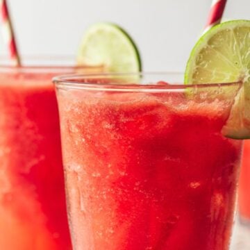 Three glasses of refreshing watermelon slushies are seen with red and white striped straws and lime slices on the rim. The drinks are bright red, garnished with lime wheels, and are placed on a light surface with a blurred background, perfect for anyone searching for simple drink recipes.