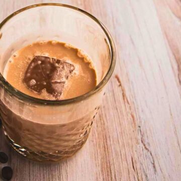 A glass of iced coffee with a large ice cube, surrounded by scattered coffee beans on a light wooden surface.