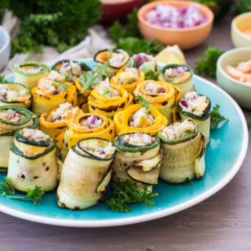 A plate of rolled zucchini and squash slices filled with a creamy mixture, garnished with parsley, is set on a table. Perfect for those seeking quick recipes, bowls of red onions, seasonings, and shrimp are visible in the background, alongside fresh parsley.