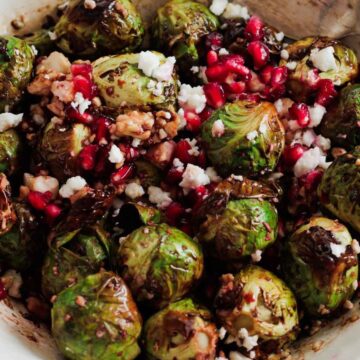 A white bowl contains roasted Brussels sprouts mixed with pomegranate seeds, crumbled feta cheese, and chopped walnuts. A silver spoon rests on the side of the bowl. The dish is set on a white wooden surface, making it perfect for those seeking quick recipes.