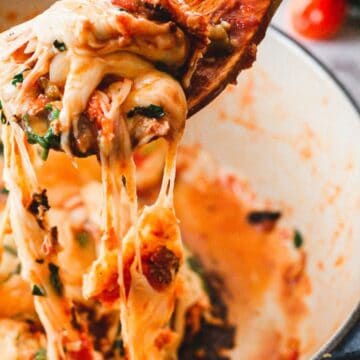 A close-up of a wooden spoon lifting a portion of cheesy, baked pasta from a white pot. The cheese is visibly stretchy, with pieces of pasta, herbs, and tomato sauce clinging to it. Fresh parsley is scattered on top, perfect for those who love quick recipes on a marble countertop background.
