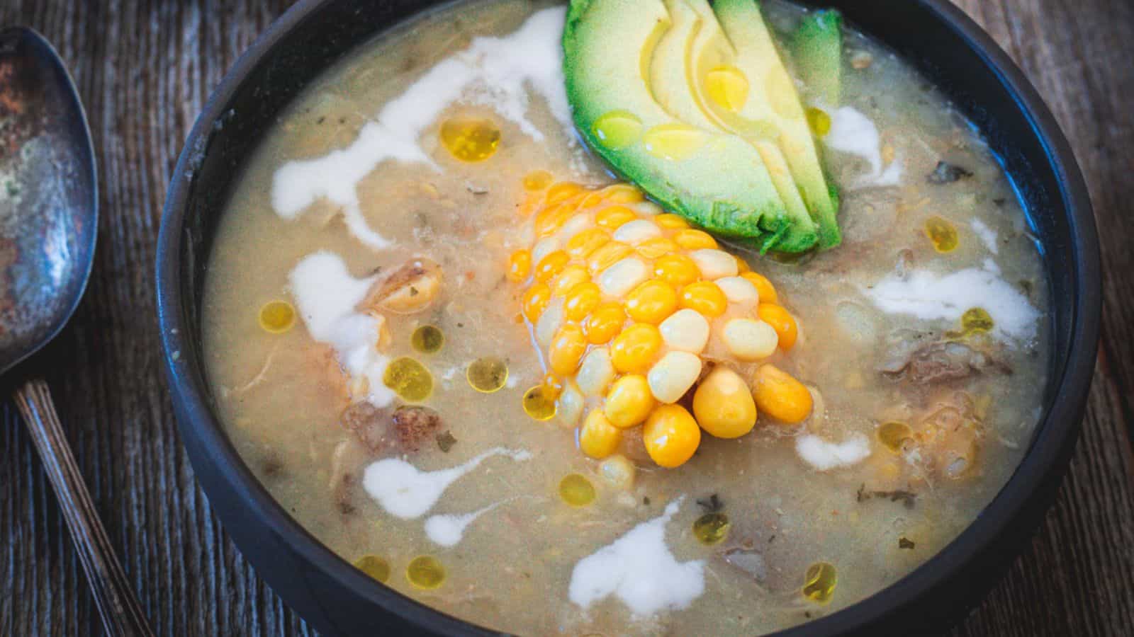 A bowl of traditional South American soup with corn, avocado, and chicken, garnished with herbs and a creamy drizzle.