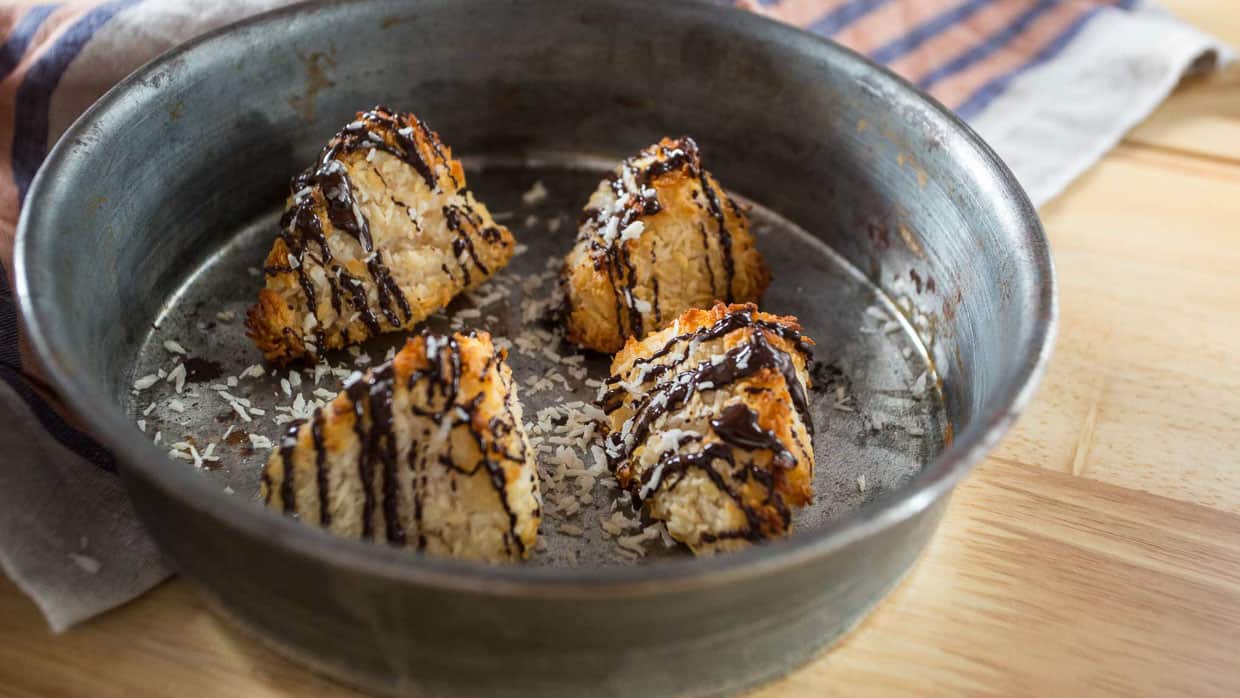 A circular metal baking pan containing four triangular coconut macaroons drizzled with chocolate is placed on a wooden surface. A striped cloth napkin is partially visible to the left of the pan. Some coconut flakes are scattered inside the pan, reminiscent of classic Passover recipes.