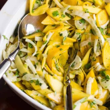 A white dish contains a salad made of yellow mango slices, fennel, and herbs. Two serving spoons are placed inside the bowl. The background is dark, which makes the vibrant colors of the salad stand out—a perfect example of quick recipes that deliver both flavor and visual appeal.