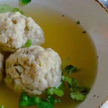 A bowl of clear soup with three large matzo balls garnished with fresh cilantro, presented on a delicately patterned white tablecloth with a spoon, embodying classic Passover recipes.