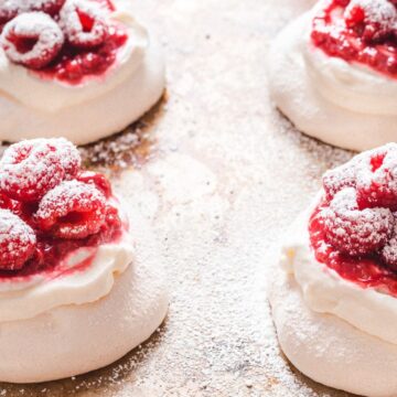 Four individual pavlovas topped with whipped cream, raspberry sauce, fresh raspberries, and a dusting of powdered sugar are arranged on a baking tray sprinkled with powdered sugar—a beautifully reimagined take on nostalgic desserts.