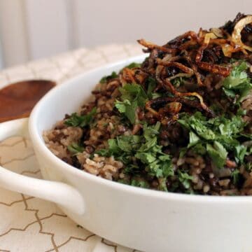 A bowl of mujadara, consisting of cooked lentils and rice, topped with crispy fried onions and chopped parsley from Grandma's recipes, served on a table. A wooden spoon rests beside the bowl