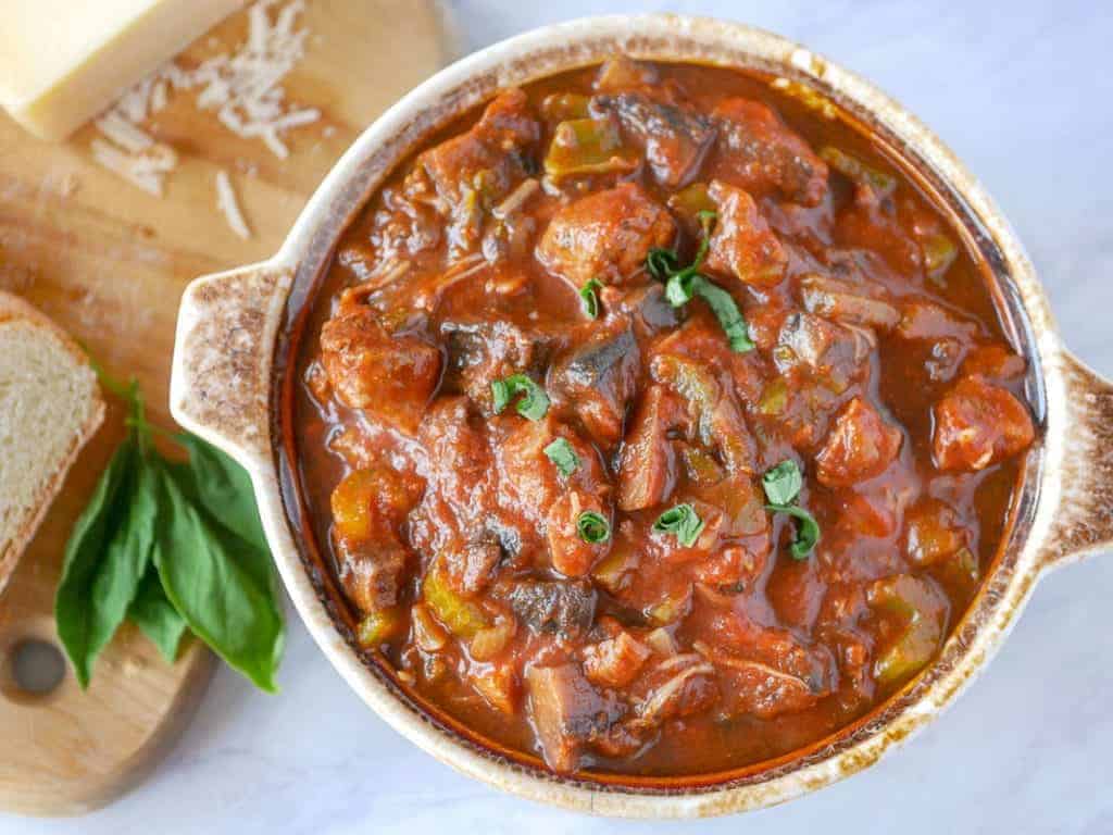 A bowl of homestyle stew garnished with fresh herbs, accompanied by a piece of bread and a block of cheese on a white background.