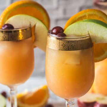Two chilled glasses of must-try fruity beverage garnished with apple slices, orange segments, and cherries, presented on a table with various fruits in the background.