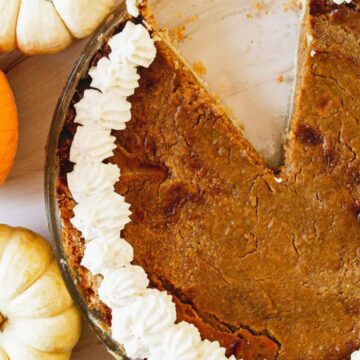 A pumpkin pie with a slice removed, garnished with whipped cream around the edge. Three small pumpkins—one orange and two white—are arranged next to the pie on a light-colored surface. Remember This? This classic dessert brings back cozy memories of autumn gatherings.
