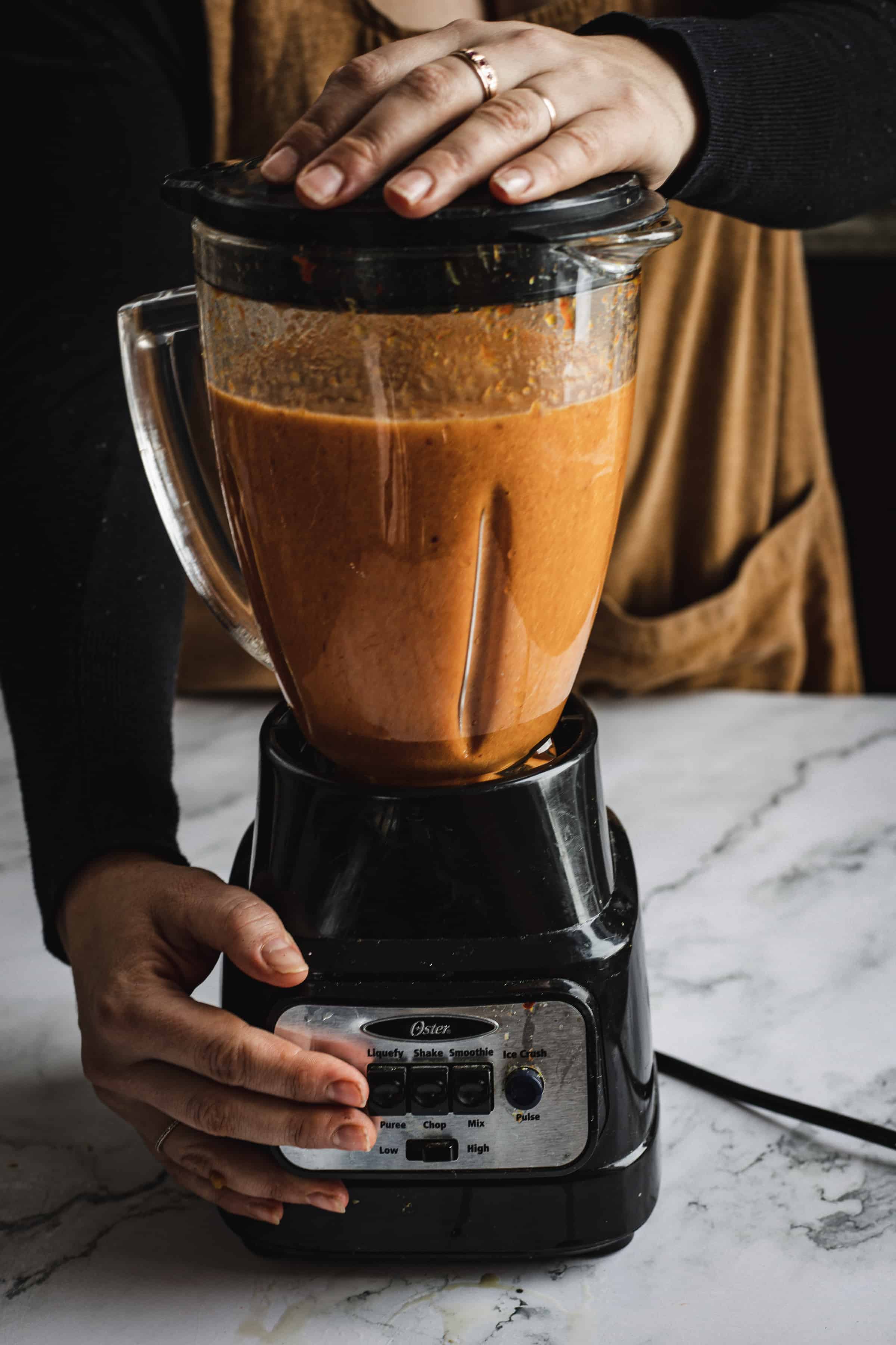 A person in an apron operating a blender filled with a vibrant bell pepper bisque on a marble countertop. Their hands are pressing the lid and selecting settings on the blender.
