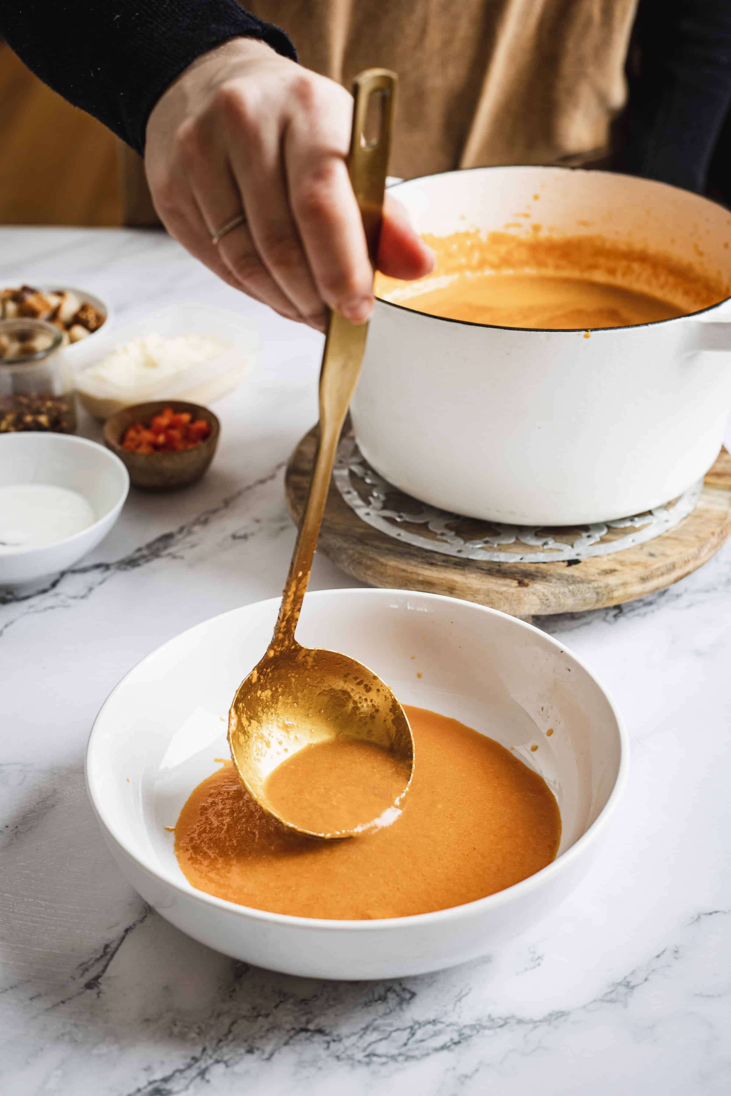 A person serving bell pepper bisque from a white pot into a bowl using a golden ladle, with ingredients like cream and spices visible on the marble countertop.
