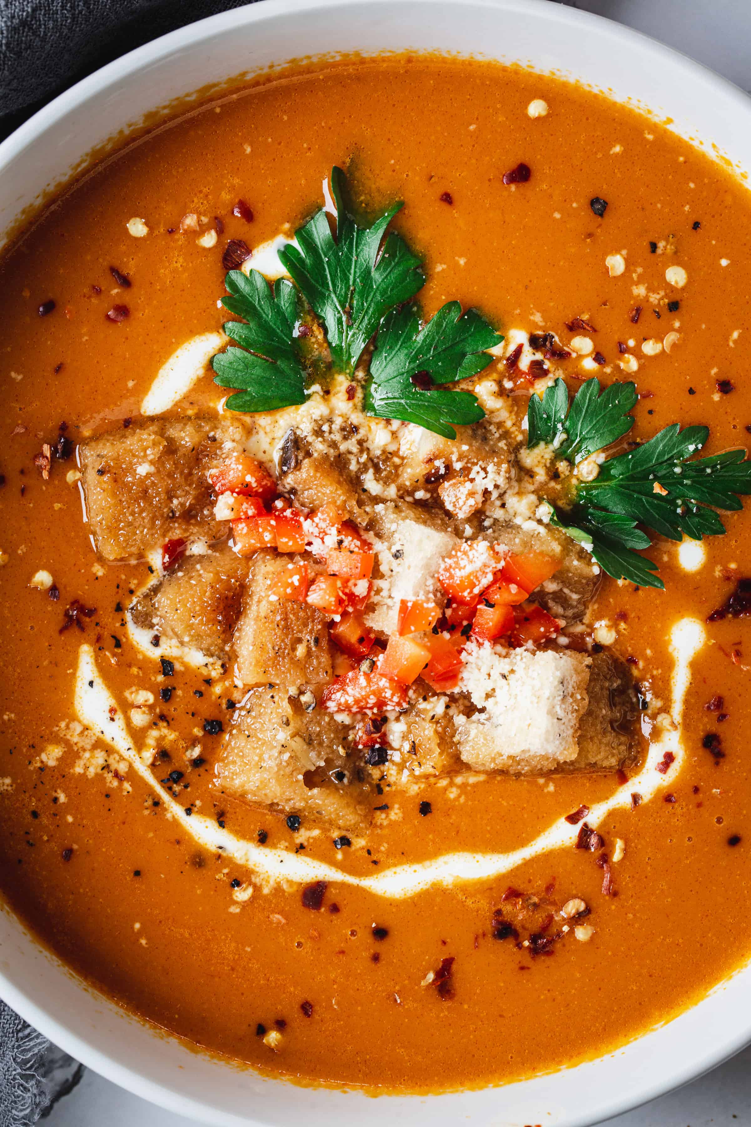A bowl of creamy tomato bisque garnished with croutons, chopped red bell peppers, fresh parsley, and a drizzle of cream, sprinkled with crushed black pepper.
