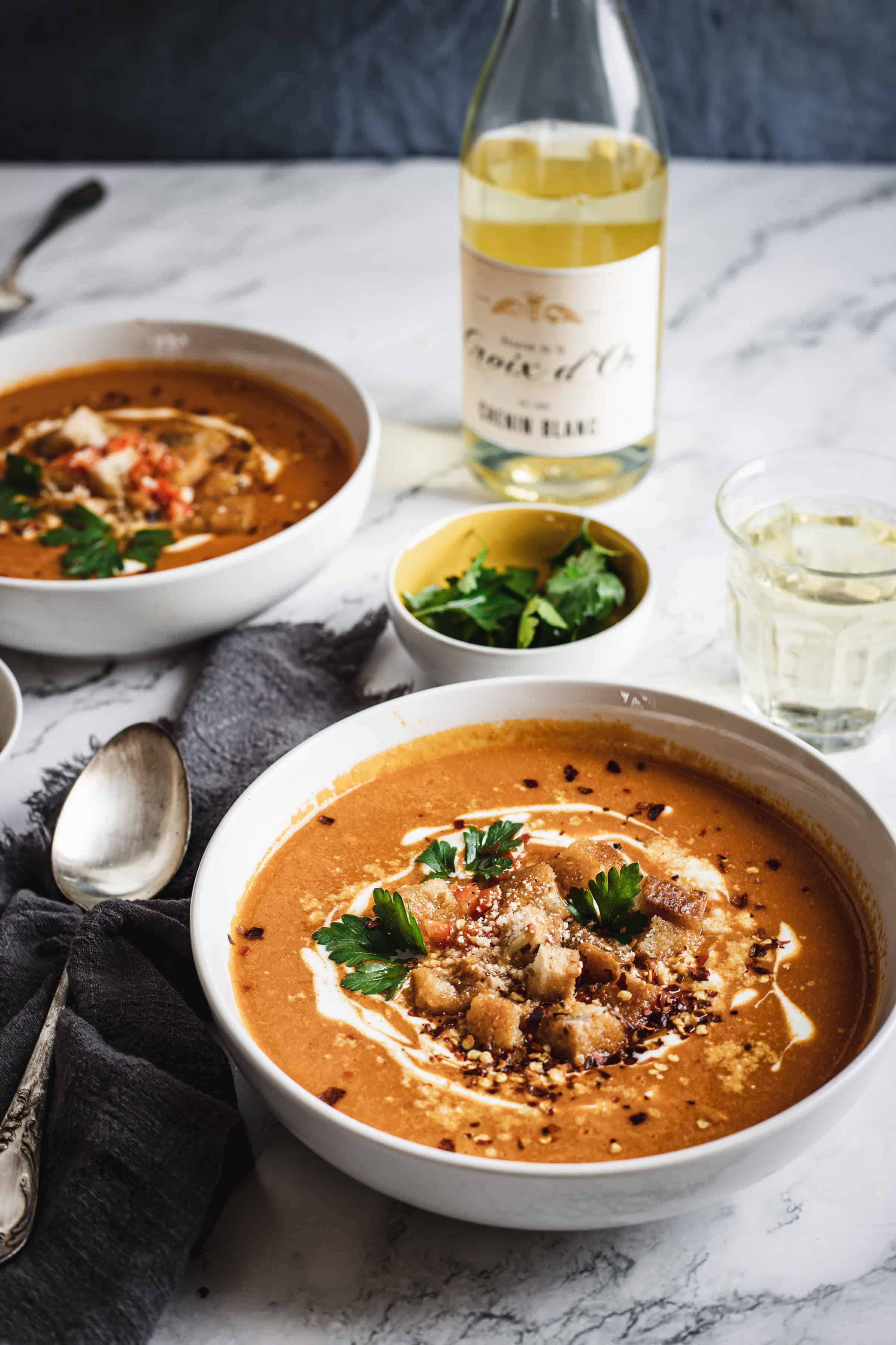 Two bowls of creamy bell pepper bisque topped with herbs and croutons, with a bottle of white wine and a glass on a marble table. A cozy, elegant meal setting.